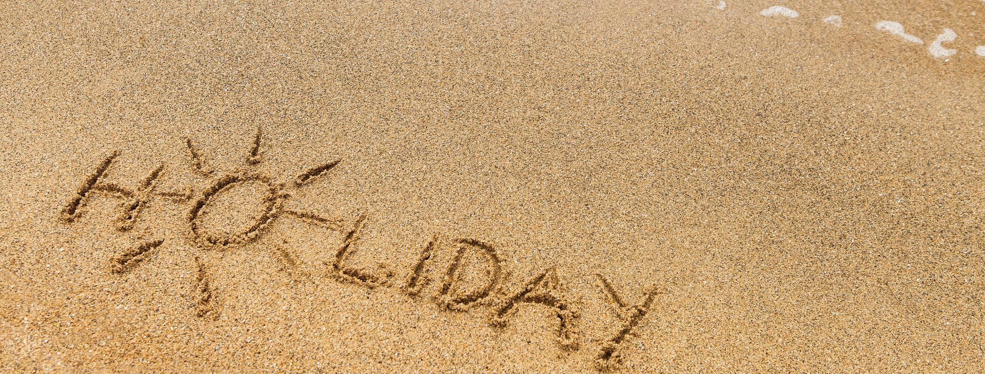 Napis 'holiday' i rysunek słońca na piasku plaży.